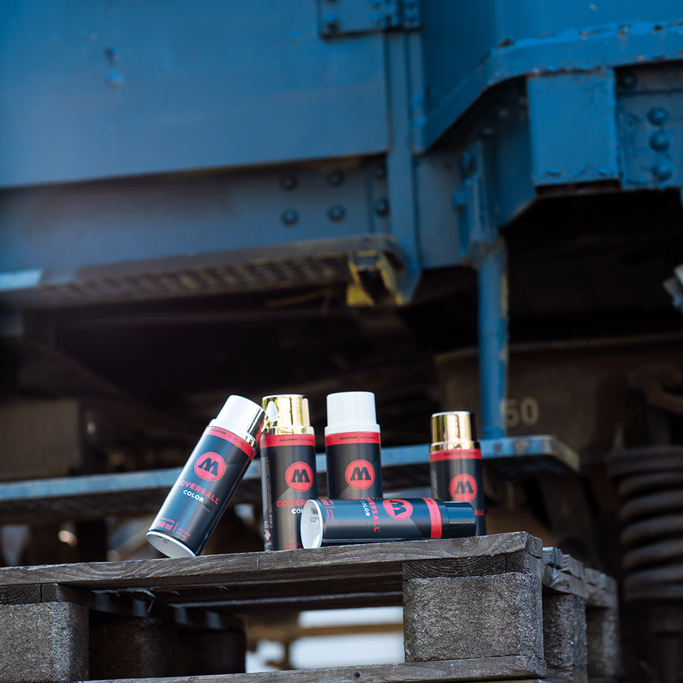 Cinque bombolette spray MOLOTOW COVERSALL Color 400ml in nero, bianco, oro e cromo su pallet di legno.